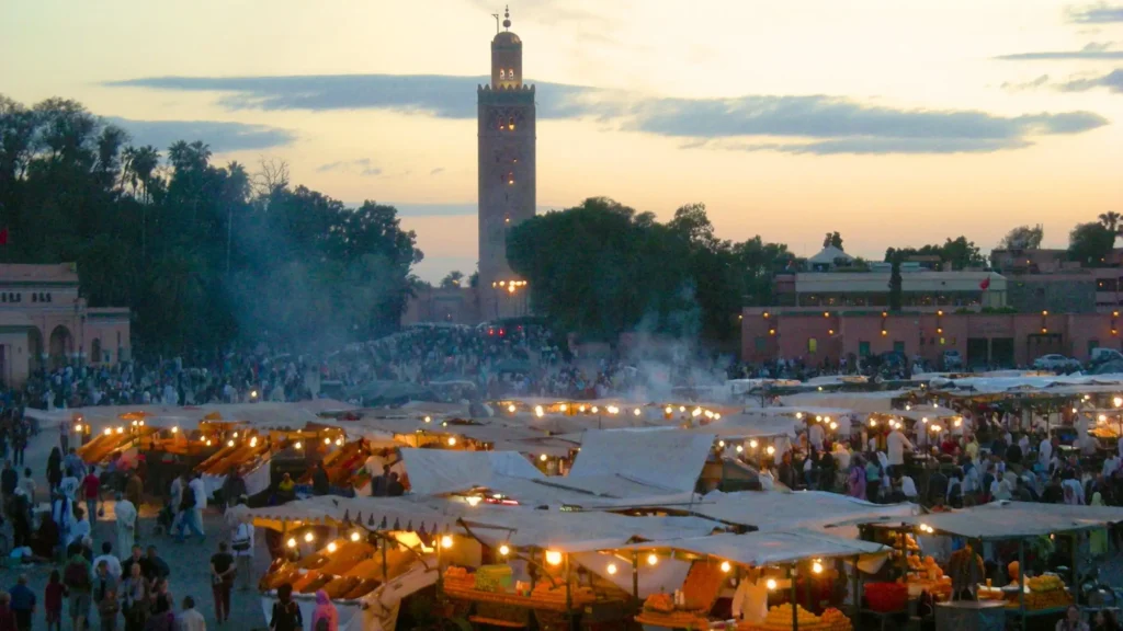 Crowded Jemaa el-Fnaa square at sunset with market stalls, lights, smoke, and the Koutoubia Mosque in the background in Marrakech, Morocco.