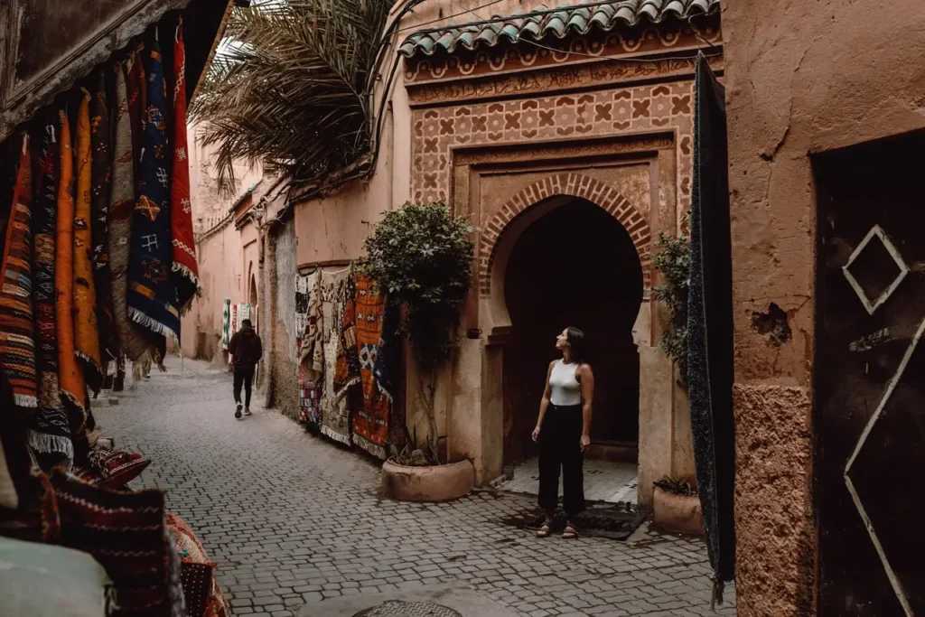 Narrow alley in the Marrakech Medina with hanging Berber carpets, traditional pink walls, an arched doorway, and a woman admiring the architecture.