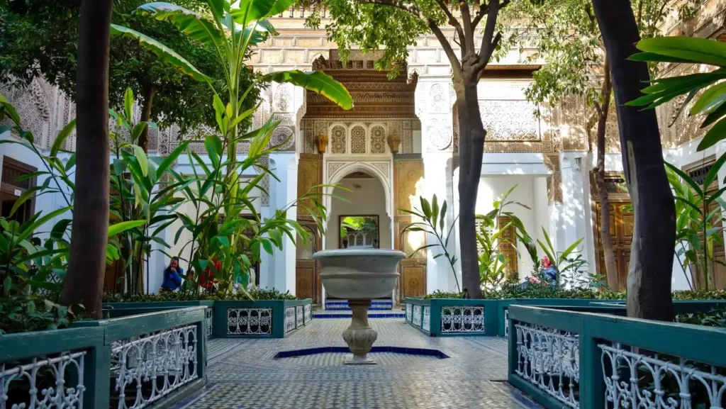 Interior courtyard of the Bahia Palace in Marrakech with a central fountain, green plants, mosaic tiles, and ornate Moroccan architectural details.