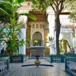 Interior courtyard of the Bahia Palace in Marrakech with a central fountain, green plants, mosaic tiles, and ornate Moroccan architectural details.