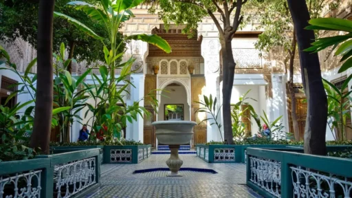 Interior courtyard of the Bahia Palace in Marrakech with a central fountain, green plants, mosaic tiles, and ornate Moroccan architectural details.