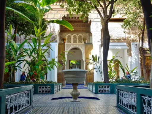 Interior courtyard of the Bahia Palace in Marrakech with a central fountain, green plants, mosaic tiles, and ornate Moroccan architectural details.