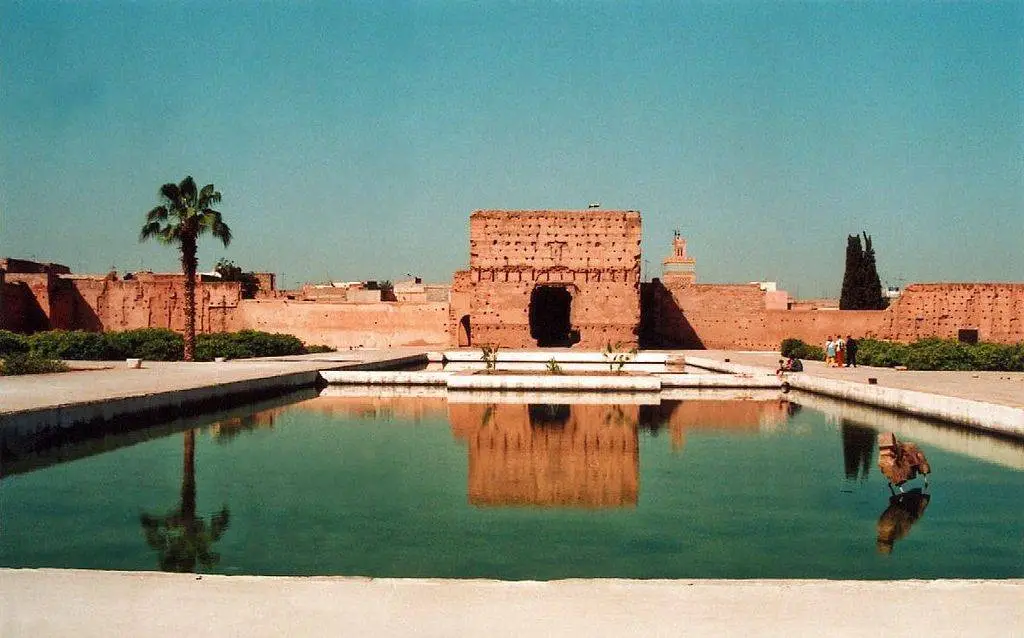 Large reflective pool in front of the historic ruins of El Badi Palace in Marrakech, with palm trees and ancient reddish walls under a clear blue sky.