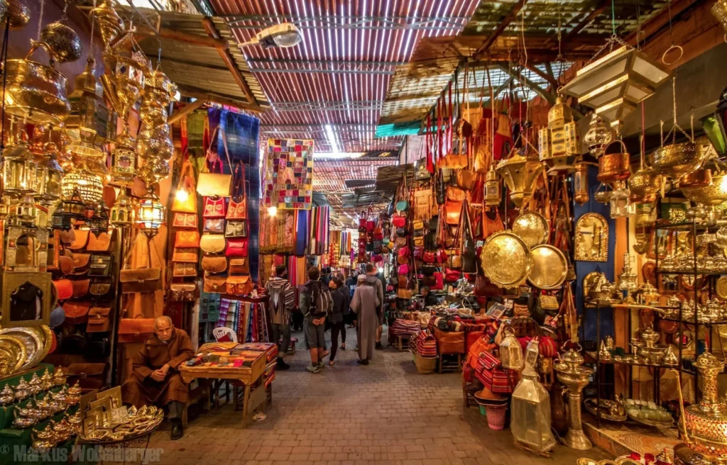 Traditional souk in Marrakech with leather bags, brass lamps, colorful textiles, and people walking through the lively market stalls.